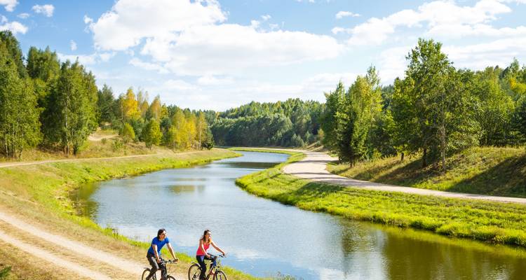 Zwei Radfahrer fahren auf einem Schotterweg neben einem sich windenden Fluss, der von Mischwald gesäumt ist, an einem sonnigen Nachmittag.