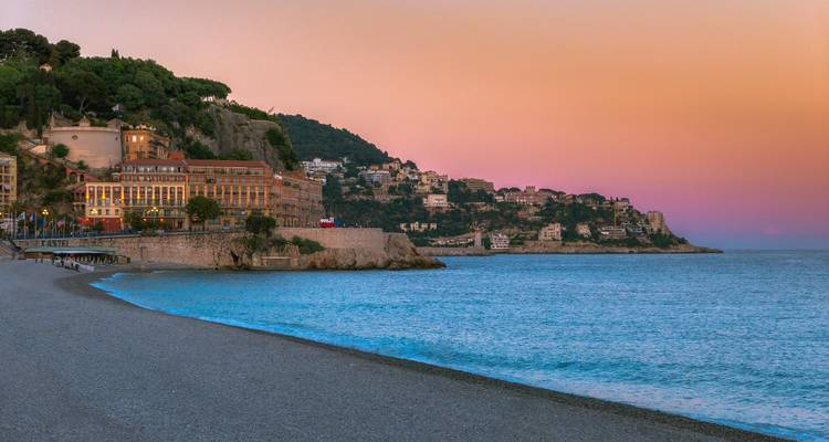Pastellrosa und orangefarbene Himmel leuchten über einem Kieselstrand und einer Küstenstadt in der Abenddämmerung.
