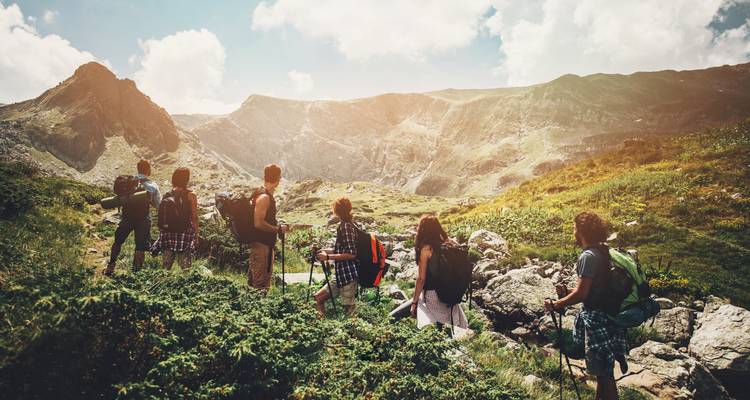 Eine Gruppe von Rucksacktouristen macht eine Pause inmitten felsiger Alpenlandschaft und blickt zu aufragenden Graten unter dramatischem Sonnenlicht empor.
