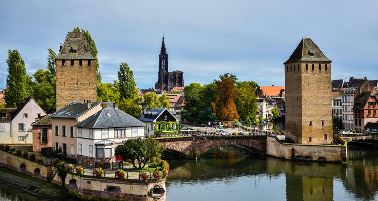 Iconische middeleeuwse torens die een rivier overspannen met een gewelfde brug en de spits van de kathedraal van Straatsburg die op de achtergrond oprijst.