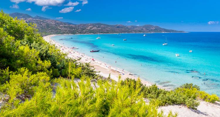 Lebendige türkisfarbene Bucht mit Sandstrand, Yachten und fernen Bergen unter einem strahlend blauen Himmel.