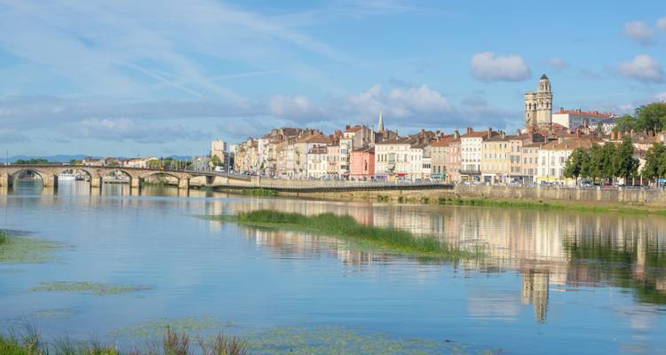 Ruhige französische Flussuferstadt mit pastellfarbenen Gebäuden und gewölbter Brücke, die sich in stillem Wasser spiegelt.