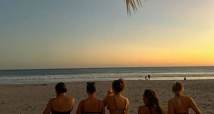 Un grupo de amigos se sienta en la arena viendo el sol ponerse sobre las olas suaves.