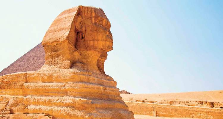 Seitenansicht der Großen Sphinx mit einer dahinter aufragenden Pyramide vor einem klaren blauen Wüstenhimmel.