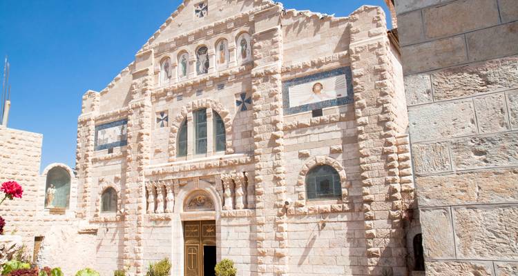 Eine cremefarbene Kirche im byzantinischen Stil mit kunstvollem Steinwerk steht vor einem klaren jordanischen Himmel.