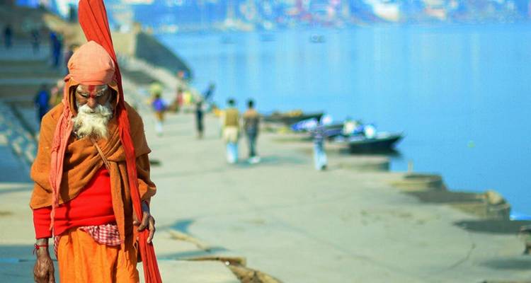 Elderly sadhu in saffron robes walks along the ghats of Varanasi beside the Ganges River.