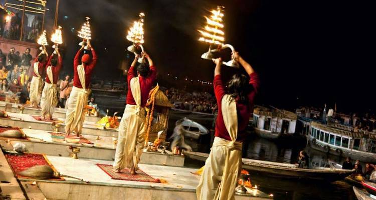 Priests perform an evening Ganga Aarti ritual holding blazing oil lamps on a riverfront platform while boats and onlookers watch nearby.