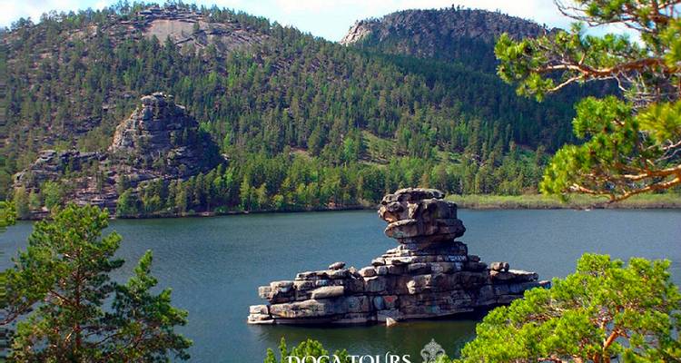 Collines couvertes de pins et un îlot rocheux en forme de champignon dans un lac de montagne transparent avec un logo de tour visible.