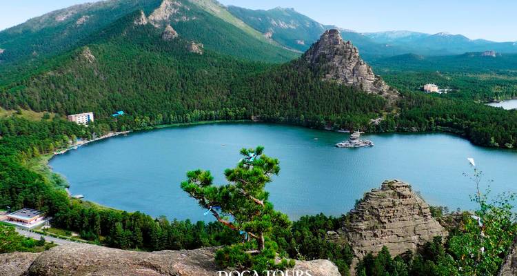 Vue en hauteur d'un lac de montagne rond et bleu entouré de pics boisés, filigrane de visite présent.