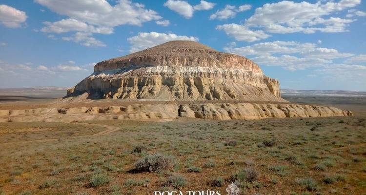 Butte isolée au sommet plat se dressant dans une large steppe semi-désertique avec des arbustes épars sous un ciel partiellement nuageux.