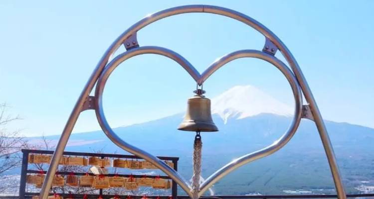 Metal heart-shaped frame with a hanging bell perfectly encircling snow-capped Mount Fuji against a clear blue sky.