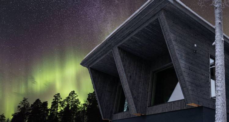Noorderlicht danst boven een slanke donkere houten hut tegen een sterrenrijke winterse nachtelijke hemel.