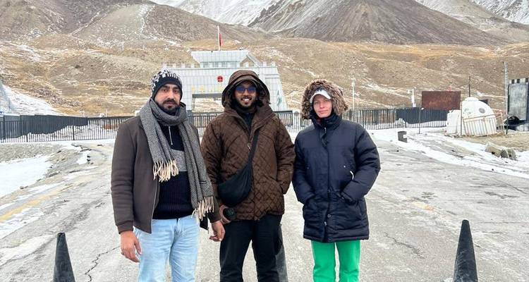 Trois hommes en vestes d'hiver se tiennent sur une route pavée de col de montagne avec des sommets enneigés et une barrière frontalière en arrière-plan.
