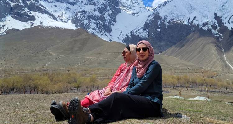 Deux femmes se détendent sur un terrain herbeux avec des pics enneigés spectaculaires et des arbres printaniers clairsemés derrière elles.