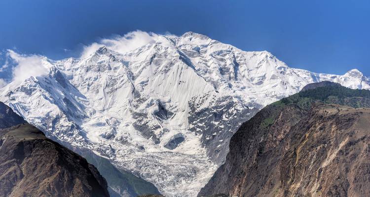Un massif montagneux imposant recouvert de neige s'élève abruptement au-dessus de gorges profondes et de crêtes boisées plus basses sous un ciel bleu dégagé.