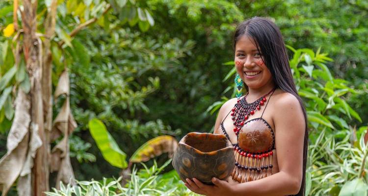 Indigenous woman smiles while holding a carved bowl, surrounded by lush jungle plants.