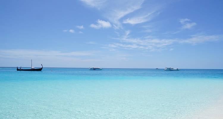Arena blanca prístina se encuentra con agua azul luminosa con pequeños botes flotando bajo un cielo tropical brillante.