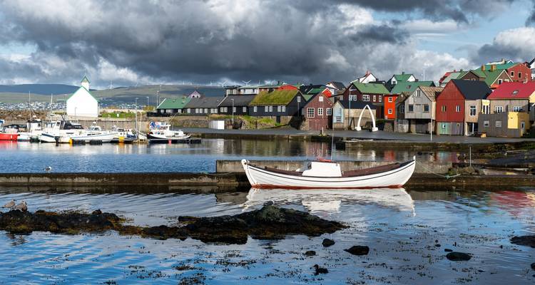 Ein kleiner Fischereihafen mit bunten grasdachgedeckten Häusern und einem weißen Boot liegt unter dramatischen dunklen Wolken.