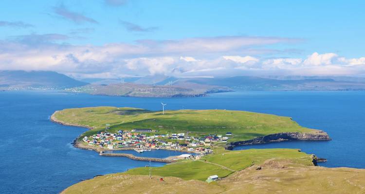 Luftaufnahme eines winzigen grünen Inseldorfes mit einer Windkraftanlage, umgeben von tiefblauem Meer und fernen sanften Hügeln.