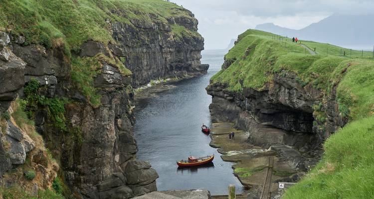Eine schmale Schlucht mit hohen grünbewachsenen Klippen öffnet sich zum Meer, wo kleine rote Boote auf dunklem Wasser treiben.