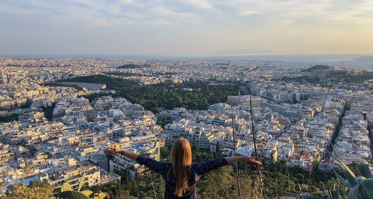 Viajero extiende los brazos en triunfo sobre una vasta vista panorámica de Atenas en la hora dorada.