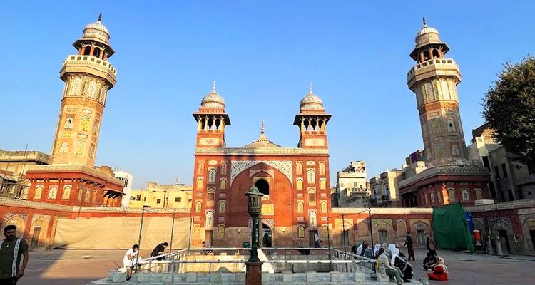 Cour de mosquée ornée en brique rouge avec trois minarets et un bassin d'ablution central sous un ciel bleu clair