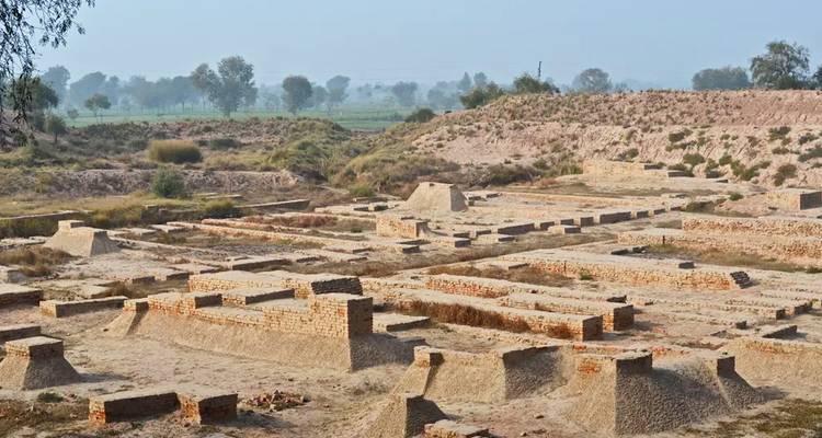 Vue large de ruines archéologiques en briques anciennes réparties à travers un paysage poussiéreux sous un ciel brumeux