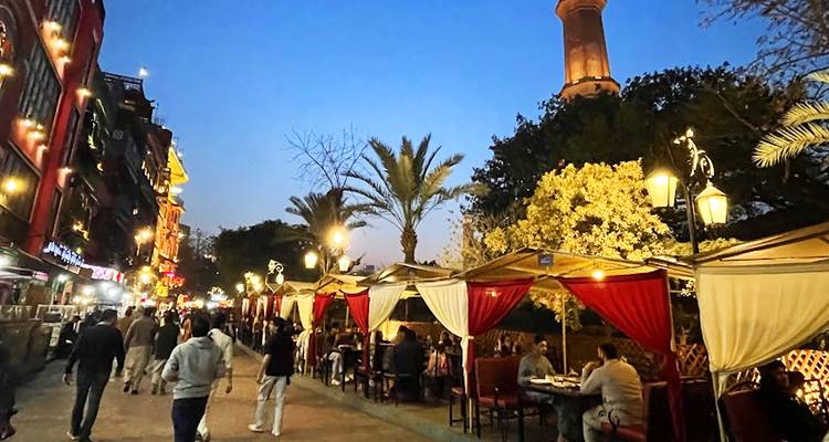 Cafés en plein air avec des tentures rouges et des clients qui dînent sous les palmiers au crépuscule avec un minaret en arrière-plan