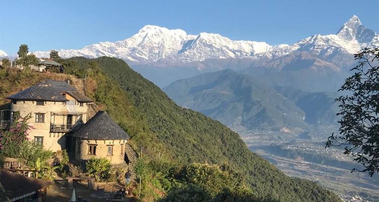 Complejo de cabañas de piedra encaramado en una cresta verde exuberante con vistas panorámicas de las cumbres nevadas del Himalaya.