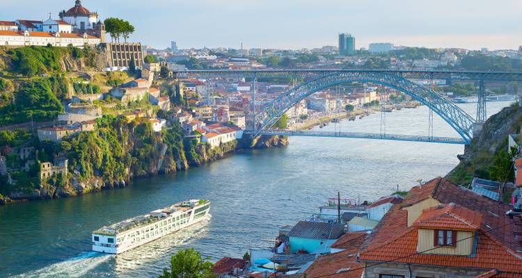Vue classique de Porto avec le pont Dom Luís I qui enjambe le fleuve Douro tandis qu'un navire de croisière blanc navigue sous les toits de tuiles en terre cuite.