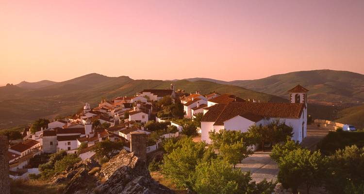 Lueur chaude du coucher de soleil sur un village perché aux murs blanchis à la chaux dans la région de l'Alentejo au Portugal, entouré de collines ondulantes.