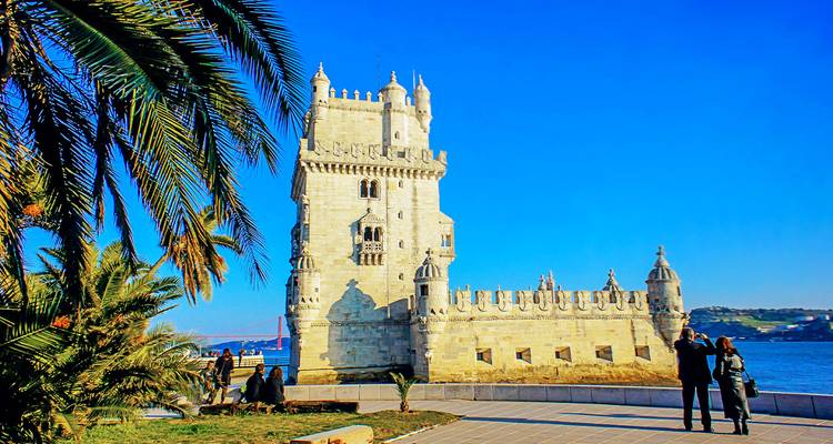 Historic Belem Tower beside the river framed by palm fronds and clear blue sky.