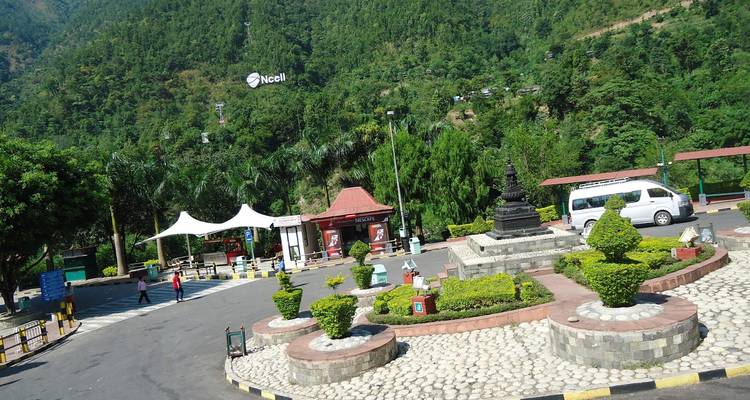 Área de descanso de montaña con jardín cuidado, cafeterías y teleférico distante en ladera boscosa