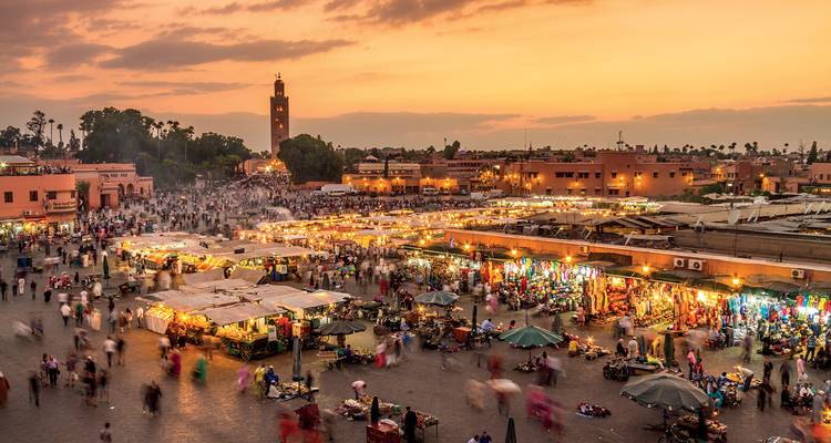 Geschäftiger Jemaa el-Fnaa-Platz in Marrakesch bei Sonnenuntergang mit Marktständen und Menschenmengen unter orangefarbenem Himmel.