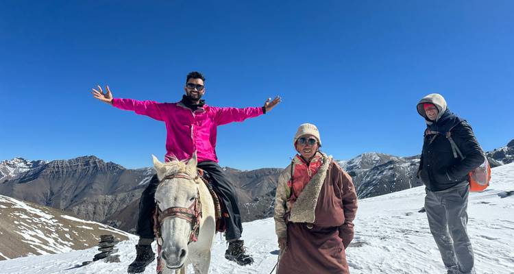 Drie trekkers, waarvan één te paard, vieren feest op een met sneeuw bedekte bergpas onder een diepblauwe hemel.