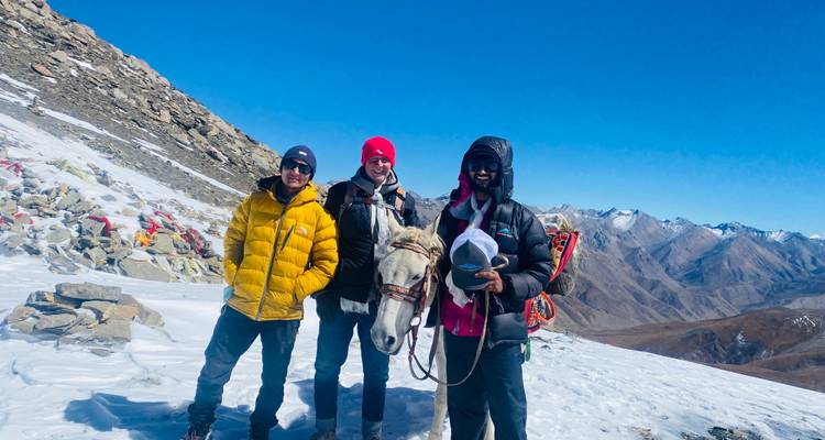 Drie trekkers met een wit paard staand op een besneeuwde bergkam met bergketens erachter.