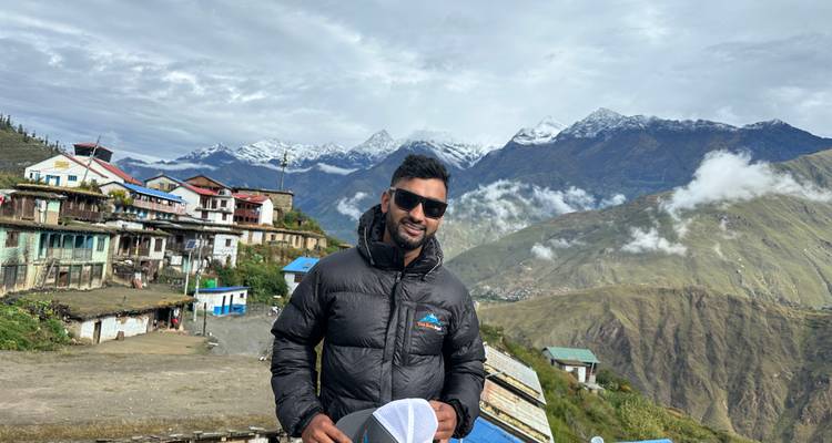 Wandelaar in zwarte jas poseert voor een bergdorp in de Himalaya met besneeuwde toppen en wolken op de achtergrond.