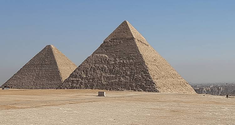 Dos pirámides masivas se alzan desde la meseta del desierto contra un cielo azul pálido con vista a El Cairo en la distancia.