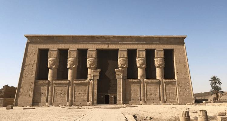 Una fachada de templo de arenisca simétrica presenta seis columnas masivas coronadas con cabezas talladas contra un cielo sin nubes.