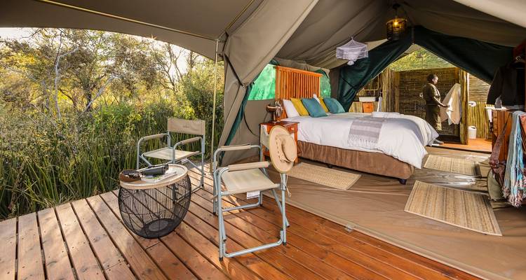 An open safari tent with wooden deck overlooks reeds while a guest prepares inside.