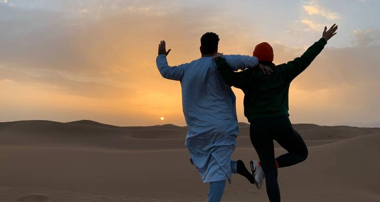 Deux amis en silhouette contre un coucher de soleil désertique, bras levés sur des dunes de sable ondulantes.