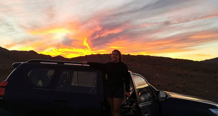 Un voyageur se tient à côté d'un VUS avec un coucher de soleil dramatique et flamboyant sur des collines rocailleuses du désert.