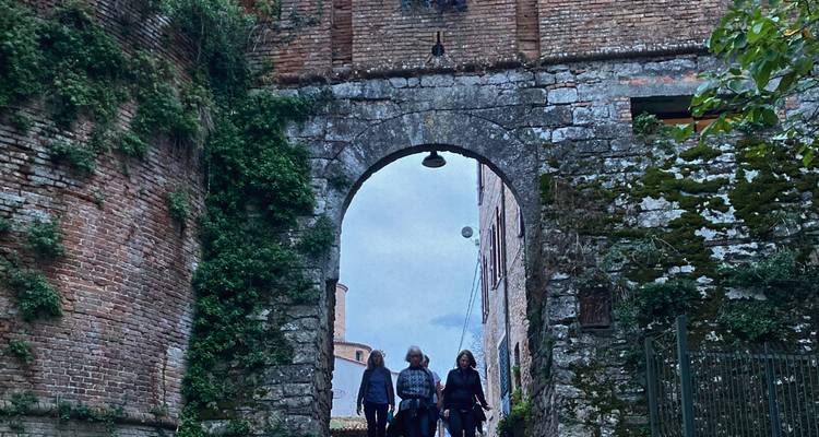 Trois voyageurs marchent sous une arche de pierre couverte de mousse et de lierre dans un village médiéval.