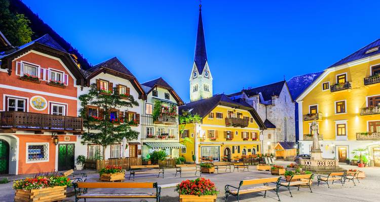 Charmanter Alpenplatz mit bunten Fassaden, Blumenkästen und einem hohen Kirchturm, beleuchtet unter einem tiefblauen Abendhimmel.