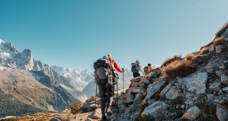 Des randonneurs équipés de gros sacs à dos et de bâtons remontent une crête alpine ensoleillée vers des sommets enneigés aux contours déchiquetés.