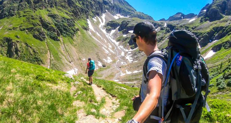Un randonneur avec une caméra d'action regarde en arrière vers un compagnon sur un sentier alpin herbeux au-dessus de neige d'été persistante.