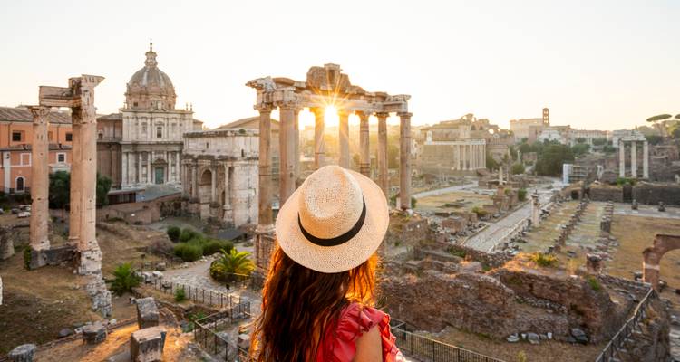 Rückansicht einer Frau mit Hut, die bei Sonnenaufgang die Ruinen des Forum Romanum bewundert, während Sonnenstrahlen durch die Säulen brechen