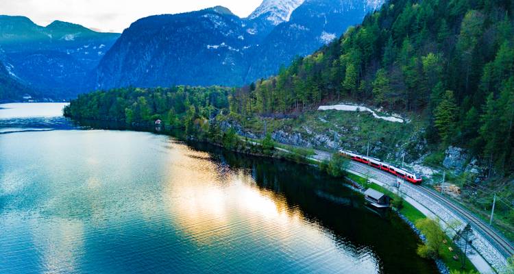 Vue aérienne d'un lac de montagne avec un train rouge circulant le long du rivage au milieu de pentes boisées et de pics enneigés