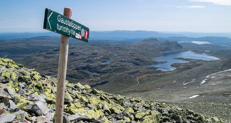 Bergtop uitzicht met een houten wegwijzer die naar Gaustatoppen wijst boven rotsachtige hellingen en meren in de verte.