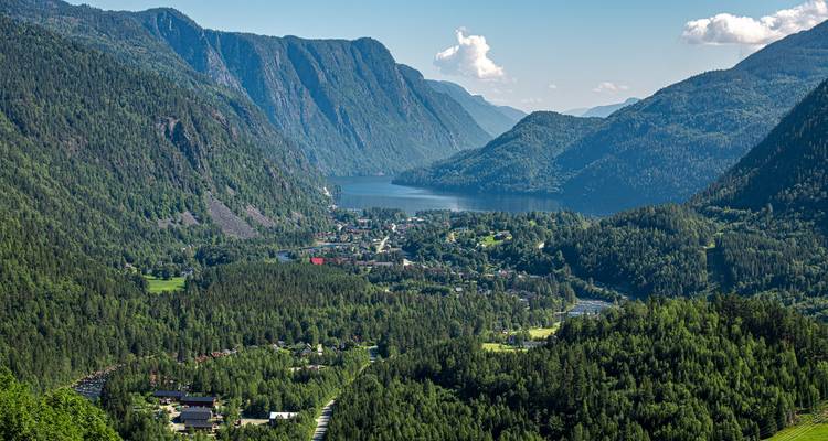 Weelderige valleistad omringd door steile blauwgroene bergen en een ver meer onder heldere luchten.